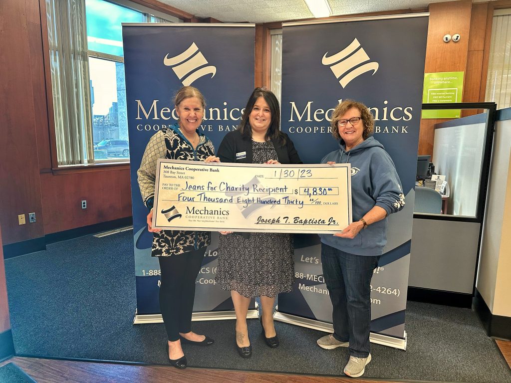 Three women stand in front of a banner with the Mechanics Cooperative Bank logo holding a large check addressed to the winners of the Employee Jeans for Charity Program
